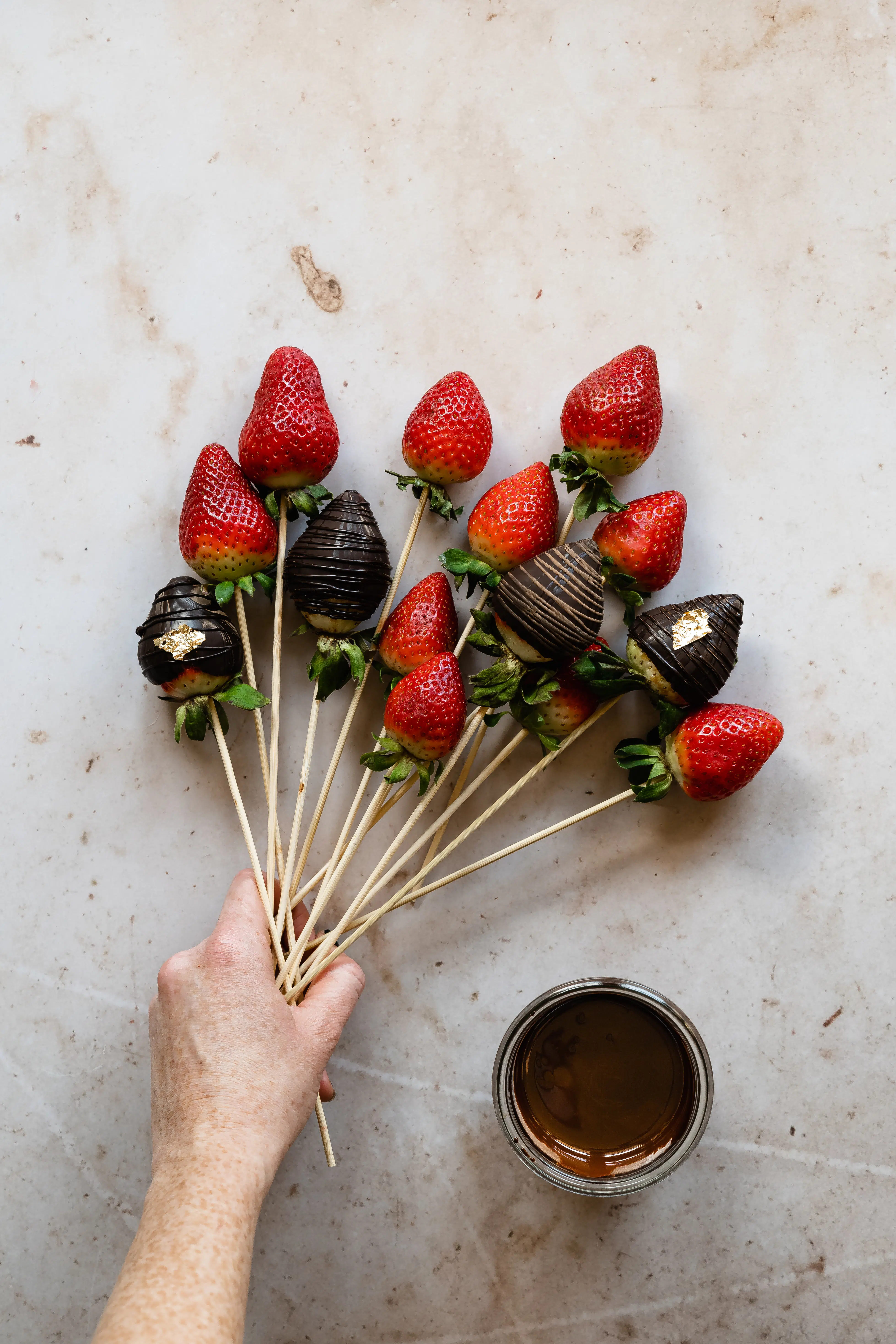 chocolate covered strawberries with bamboo skewers being held by a hand.