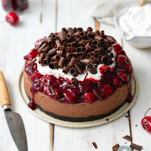 chocolate cheesecake with cherries, cream and chocolate curls on a white wooden surface.