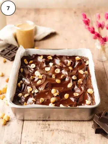 no bake brownies in a tin with ganache and toasted hazelnuts on top and a small jar of hazelnut butter in the background.