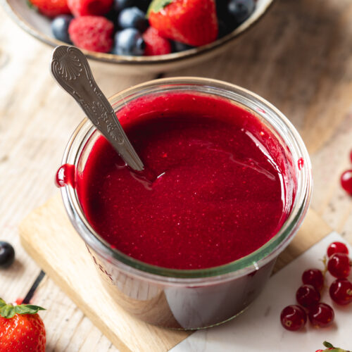 45 degree angle of a glass jar of mixed berry coulis on a wooden table with currants, strawberries, and blueberries scattered about.