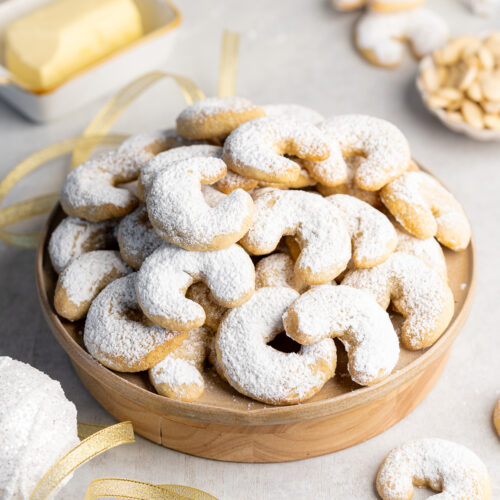 a stack of vanilla crescent cookies on a ceramic plate with a butter dish and a bowl of blanched almonds in the background.