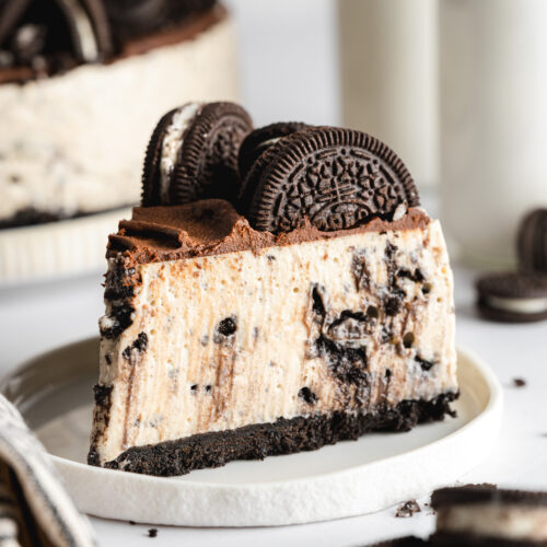 a sliced of vegan oreo cheesecake with chocolate ganache topping and oreo cookie halves on top of a ceramic white plate.