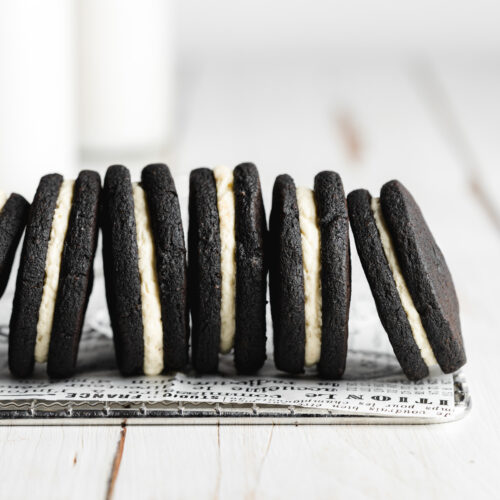 a row of homemade oreos stacked against each other on a wire rack with bottles of milk in the background.