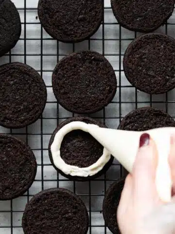 piping creme filling onto dark chocolate cookies on a wire rack.